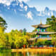 Traditional Chinese pagoda with upturned eaves reflected in river, surrounded by lush green trees with snow-capped mountains 